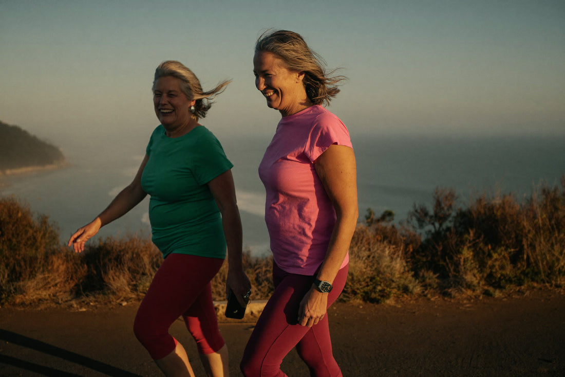 Two women in their 50s walking and laughing on a cliff-top path at golden hour, ocean behind them