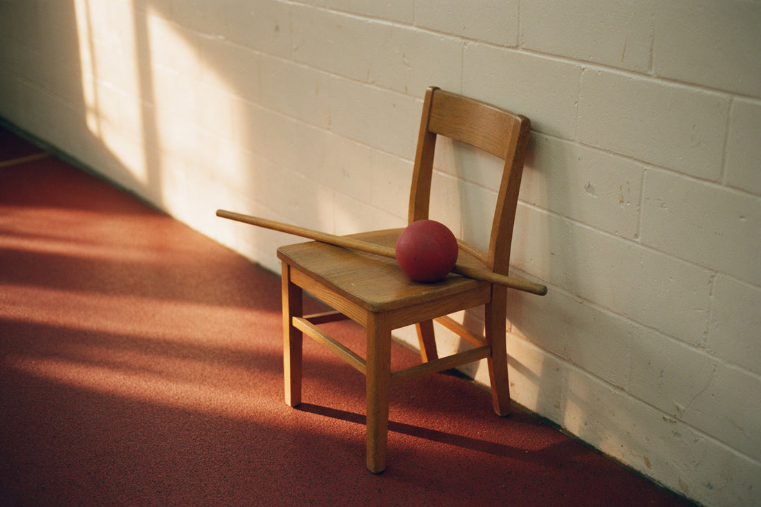 A plain wooden chair side-on to a painted wall with a broomstick resting across its seat and a small rubber ball beside it, on a rubber gym floor in afternoon light